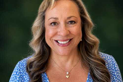 Portrait of a smiling woman with wavy light-brown hair wearing a blue patterned blouse, outdoors with a green background.