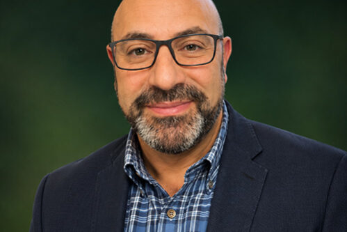 Ammar Portrait of a smiling bald man with glasses, wearing a navy blazer and blue plaid shirt, outdoors with greenery in the background.