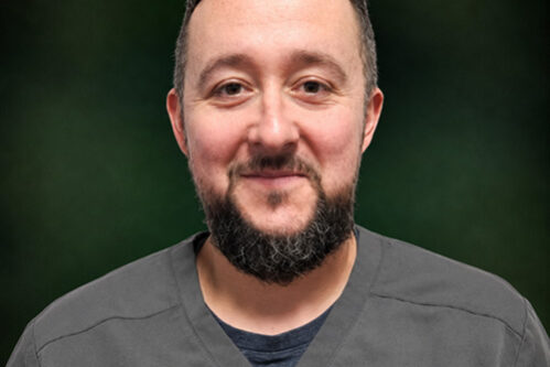 Portrait of a smiling man in gray scrubs against a dark green backdrop, likely a healthcare professional.