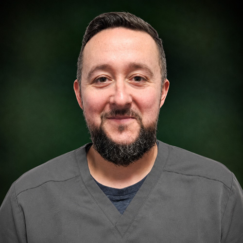Portrait of a smiling man in gray scrubs against a dark green backdrop, likely a healthcare professional.