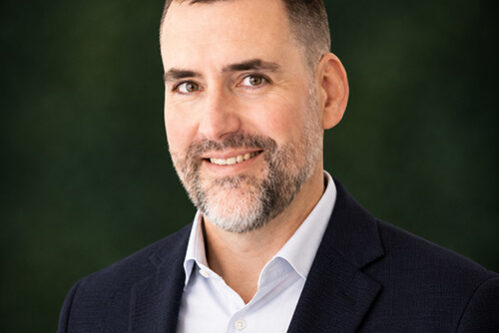 Professional headshot: smiling man in a navy blazer and light blue shirt, dark green background