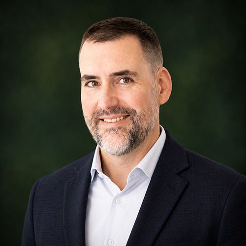 Professional headshot: smiling man in a navy blazer and light blue shirt, dark green background
