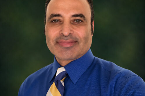 Portrait of a smiling man in a blue shirt and striped yellow, white, and blue tie, against a green background.