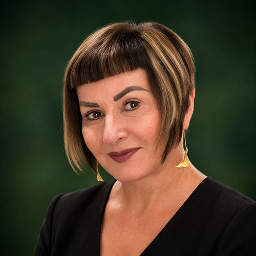 Donna Portrait of a woman with short dark hair and blonde highlights, wearing a black top and gold earrings, against a green background.