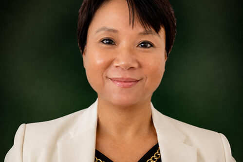Portrait of a smiling woman in a white blazer and black top with a gold chain necklace, against a dark green background?