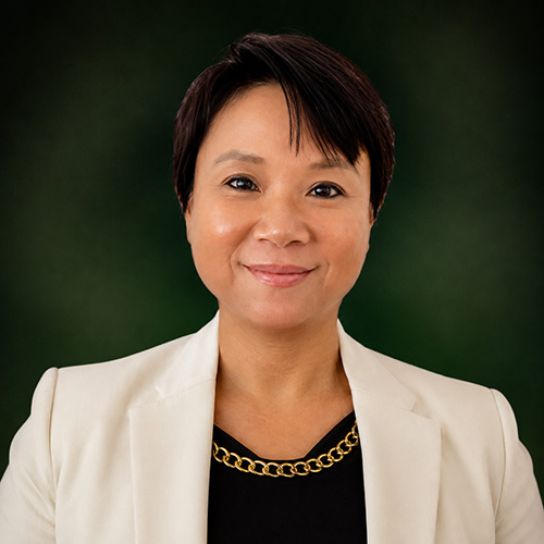 Portrait of a smiling woman in a white blazer and black top with a gold chain necklace, against a dark green background?