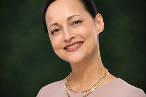 Jennifer Head-and-shoulders portrait of a smiling woman in a light pink blouse with a gold chain necklace against a dark green blurred background.