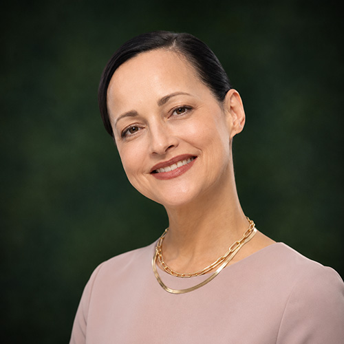 Jennifer Head-and-shoulders portrait of a smiling woman in a light pink blouse with a gold chain necklace against a dark green blurred background.