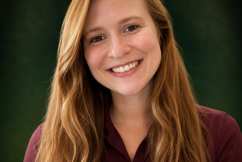 Julie Portrait of a smiling woman with long wavy red hair, wearing a maroon button-up shirt, outdoors with green background.