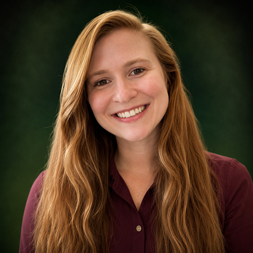 Julie Portrait of a smiling woman with long wavy red hair, wearing a maroon button-up shirt, outdoors with green background.