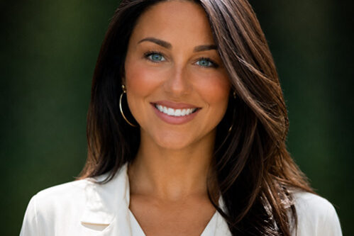 Professional headshot: smiling woman with long dark hair in a white blazer outdoors with greenery in the background.