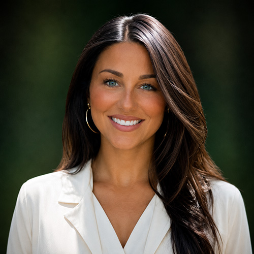 Professional headshot: smiling woman with long dark hair in a white blazer outdoors with greenery in the background.
