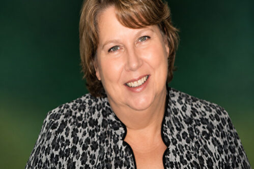 Portrait of a smiling woman with short light brown hair, wearing a black top and leopard-print jacket, against a green background.