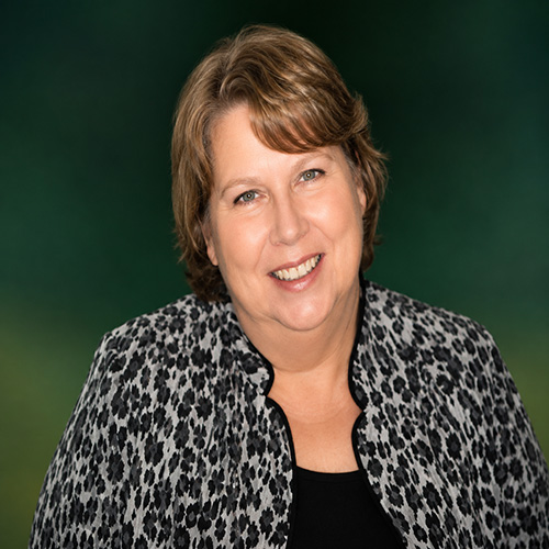 Portrait of a smiling woman with short light brown hair, wearing a black top and leopard-print jacket, against a green background.