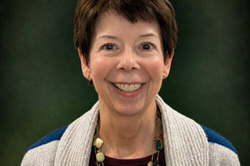 Smiling woman with short brown hair in a maroon top and striped cardigan wearing a beaded necklace, portrait against a dark green background.