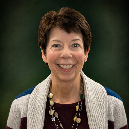 Smiling woman with short brown hair in a maroon top and striped cardigan wearing a beaded necklace, portrait against a dark green background.