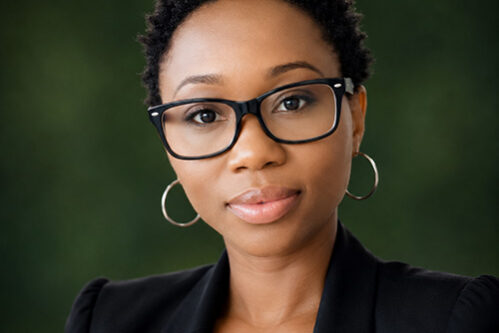 Ruby Professional woman with short curly hair, wearing a black blazer and glasses, looking at the camera with a neutral expression, green blurred background.