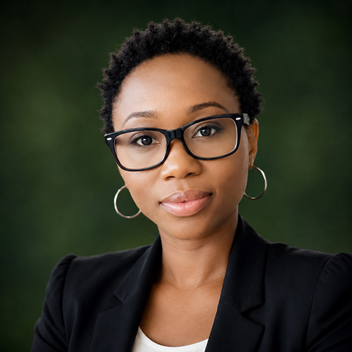 Ruby Professional woman with short curly hair, wearing a black blazer and glasses, looking at the camera with a neutral expression, green blurred background.
