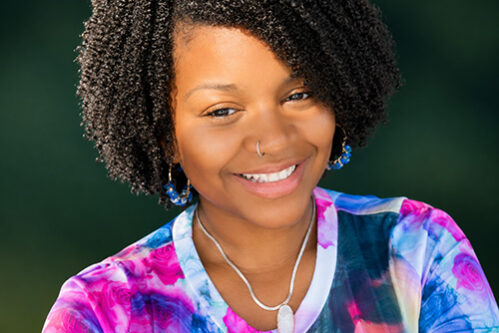 Portrait of a smiling young Black woman with curly hair, wearing a nose ring and a colorful tie-dye shirt outdoors.