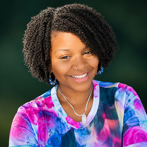 Portrait of a smiling young Black woman with curly hair, wearing a nose ring and a colorful tie-dye shirt outdoors.